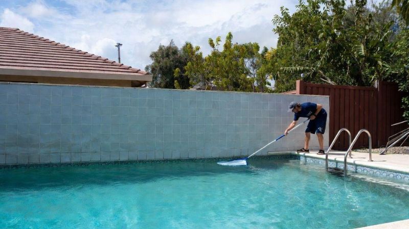 A person cleaning a swimming pool using a long pool net in a backyard area