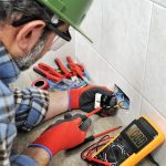 Electrician wearing gloves and a hard hat connects wires inside a wall electrical outlet using a screwdriver and pliers, with a digital multimeter nearby.