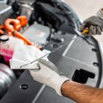 Mechanic wearing gloves inspecting the engine bay while holding a paper towel and a tool.