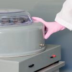 Gloved hand in a lab coat closing the lid of a gray centrifuge on a benchtop.