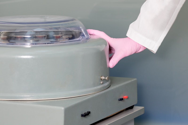 Medical Equipment and Sample Processing Gloved hand in a lab coat closing the lid of a gray centrifuge on a benchtop.