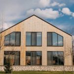 Two-story wooden house with large glass windows and a stone foundation, in a landscaped yard with leafless trees.