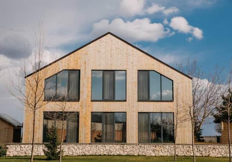 Two-story wooden house with large glass windows and a stone foundation, in a landscaped yard with leafless trees.