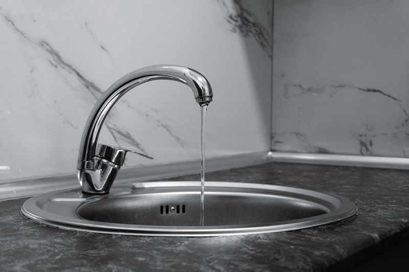 Chrome kitchen faucet pouring a thin stream into a round stainless steel sink on a dark countertop with marble backsplash (monochrome). In-use image showing running water into a basin.