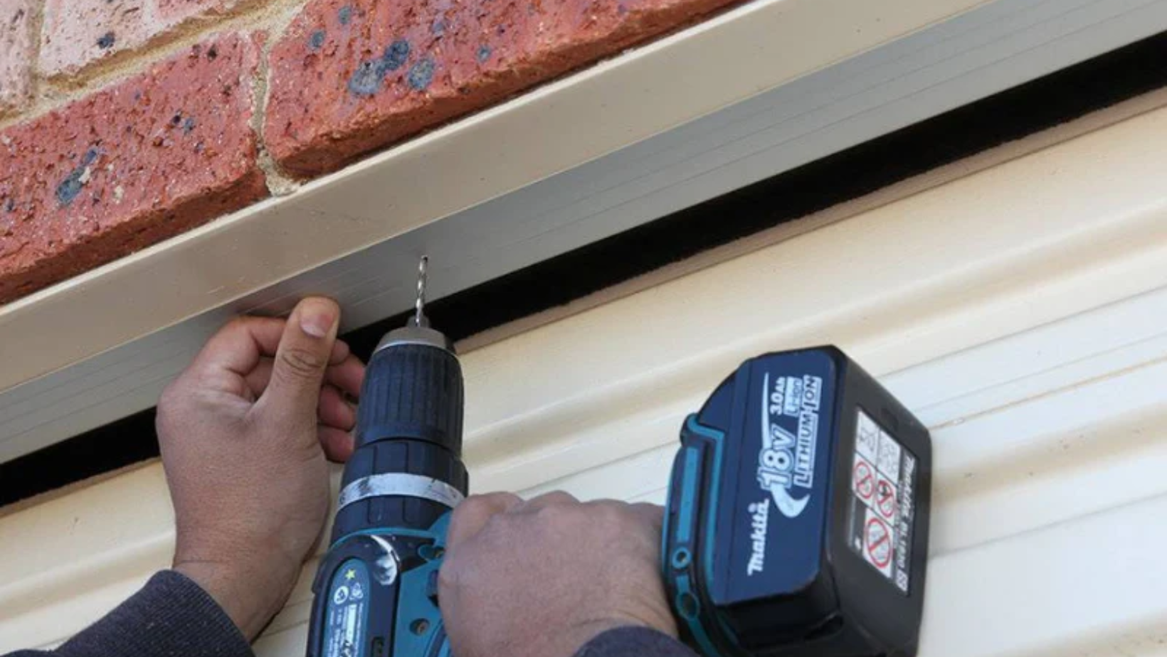 Hands wield a blue cordless drill (Makita 18V) drilling into a metal strip beneath a brick wall.