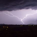 Lightning bolt divides the night sky over a dark hillside with scattered village lights below during a storm momentarily illuminating the landscape.