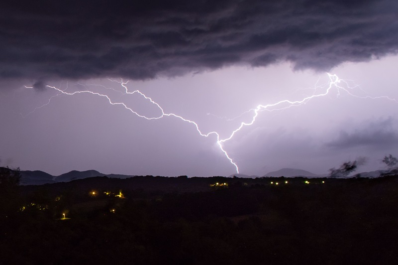 Lightning bolt divides the night sky over a dark hillside with scattered village lights below during a storm momentarily illuminating the landscape.