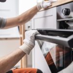 Male technician wearing safety glasses and gloves installs or removes a built-in oven door in a kitchen.