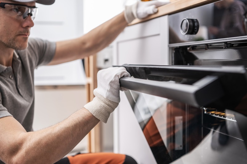 Male technician wearing safety glasses and gloves installs or removes a built-in oven door in a kitchen.