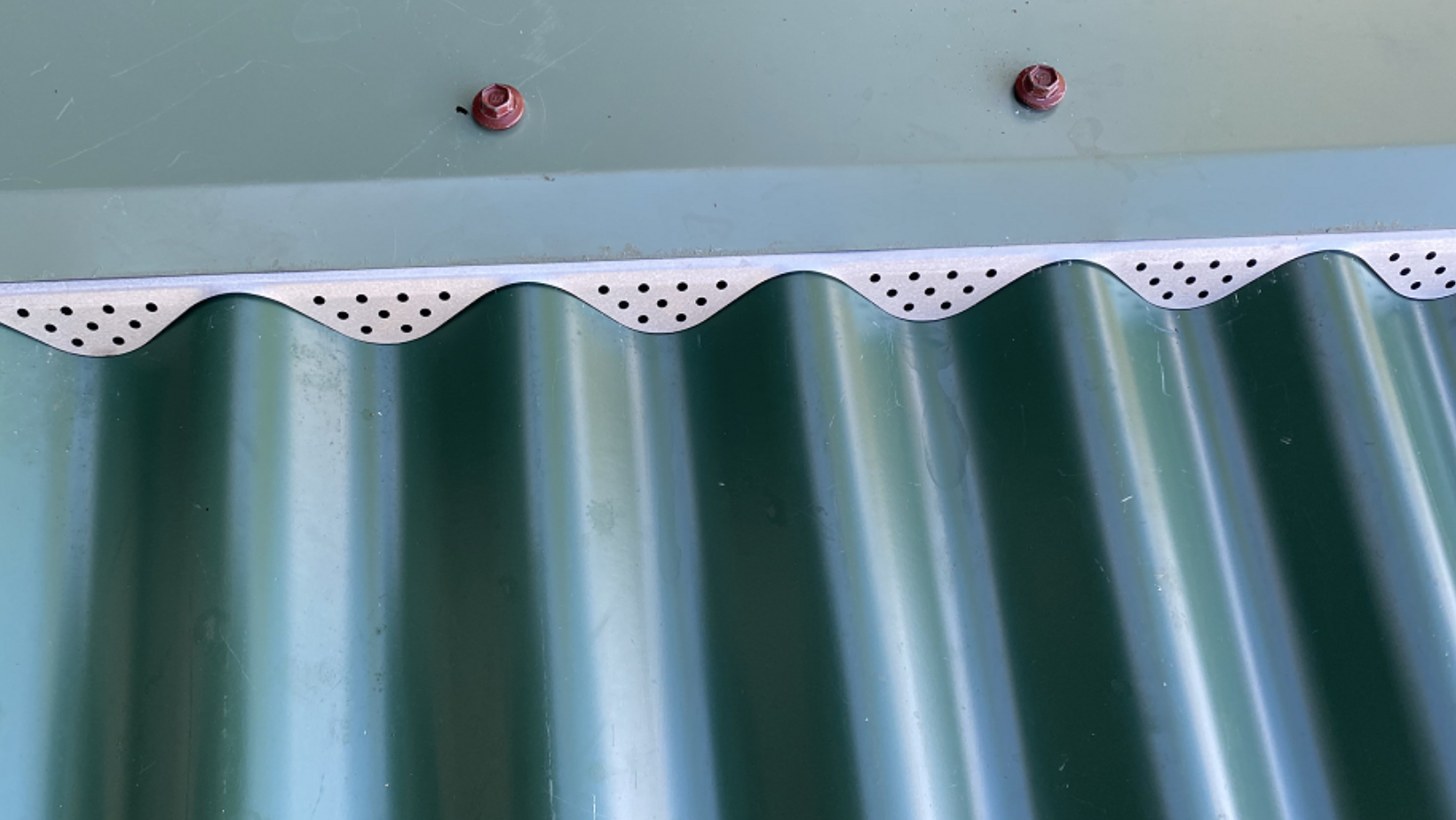 Close-up of green corrugated metal roof with perforated vented edge trim along the seam and two red bolts above it.