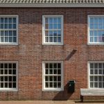 Brick building facade with six white-framed sash windows (three on each floor), a green mailbox, and a wooden bench in front.