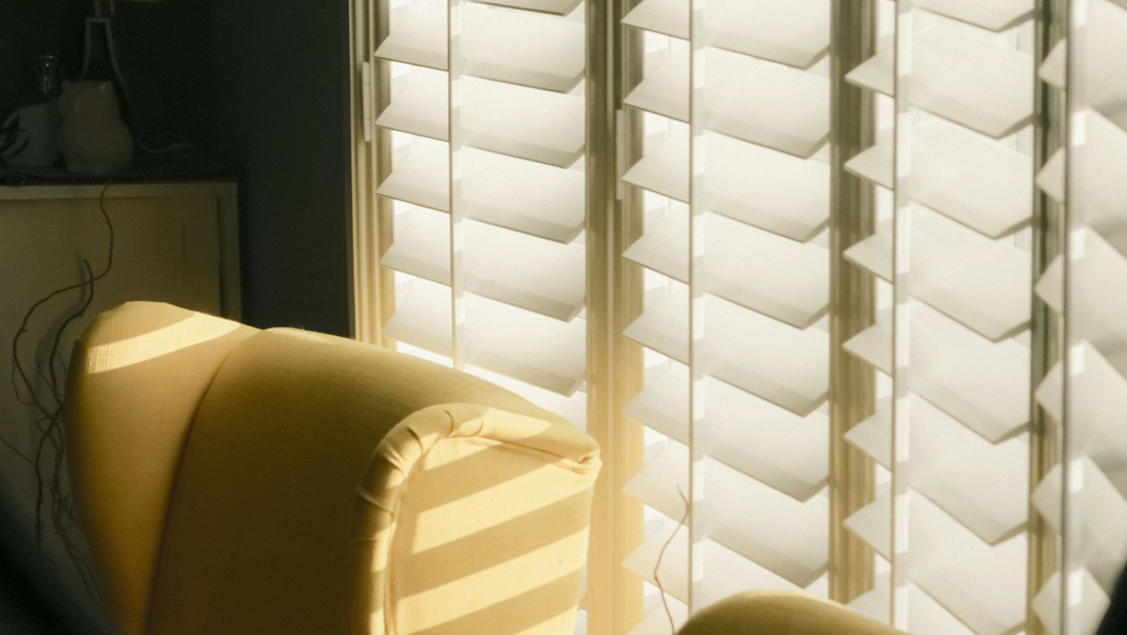 Sunlit yellow striped chair bathed in light as white plantation shutters cast shadows nearby.