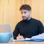 Focused businessman with beard working on a laptop, reviewing documents in a modern workspace