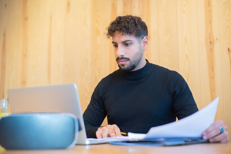 Focused businessman with beard working on a laptop, reviewing documents in a modern workspace