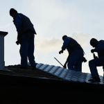 Three roofers in blue uniforms work on a metal tile roof beside a chimney, silhouetted against the sky.