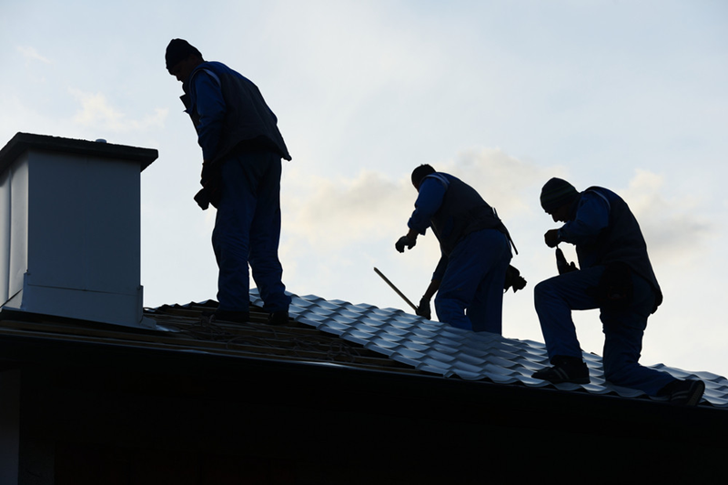 Professional Roofers Working on a Residential Home Three roofers in blue uniforms work on a metal tile roof beside a chimney, silhouetted against the sky.