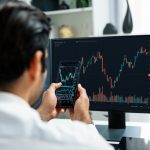 Person at a desk viewing stock charts on a large monitor while holding a smartphone with a similar chart display.
