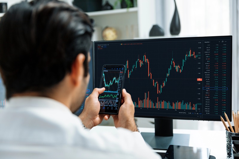 Person at a desk viewing stock charts on a large monitor while holding a smartphone with a similar chart display.