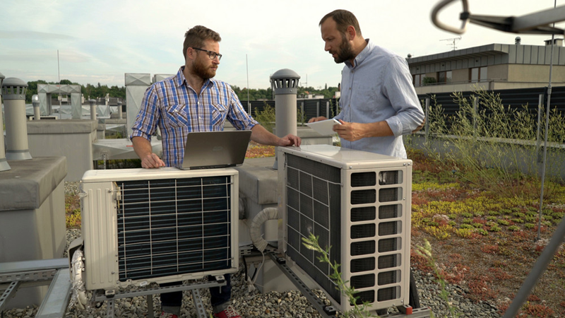 Two technicians on a building rooftop inspect rooftop HVAC units, one with a laptop and the other holding papers.