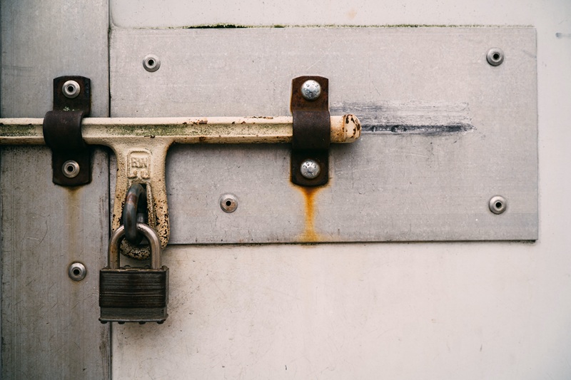 Metal door secured with a horizontal bar and padlock, showing rust and bolts on a weathered surface.