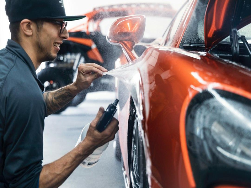 A detailer spraying solution onto the paint of an orange car in a detailing studio