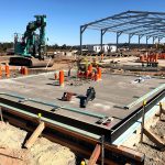 A steel workshop shed under construction, showing the concrete slab, exposed framing, and electrical conduit planning