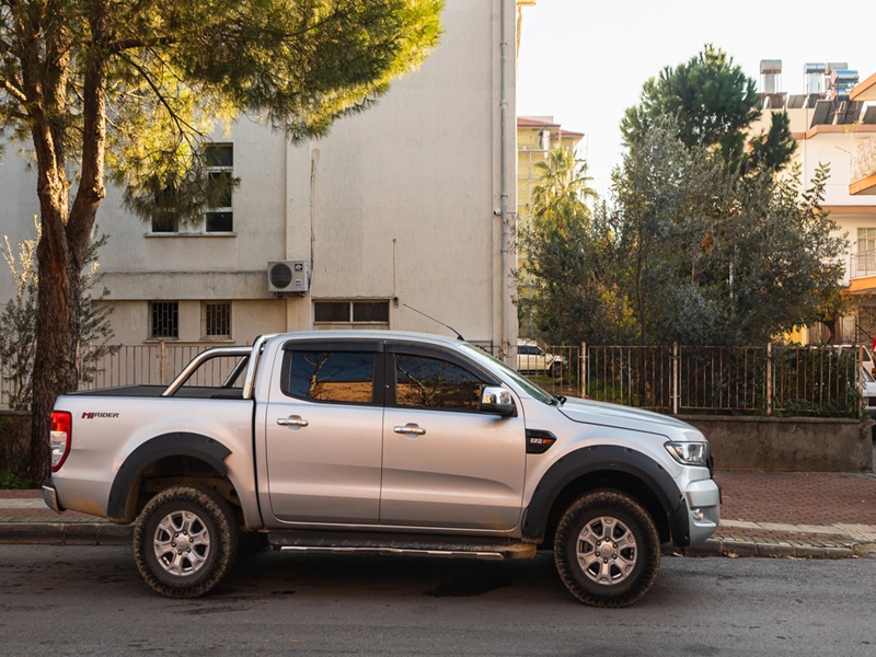 silver pickup Ford Ranger is parked on the street on a warm day against the backdrop of a fence, garden
